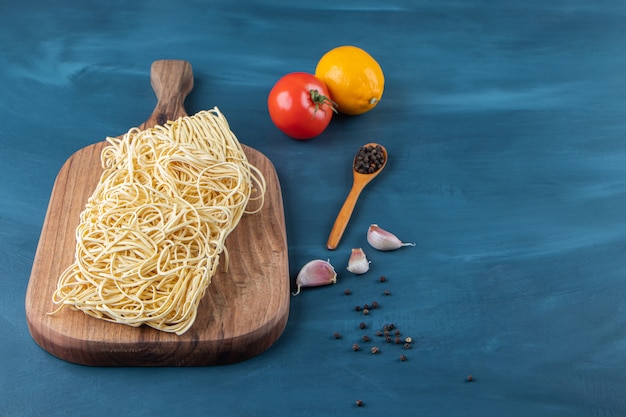 Una tabla de madera de fideos crudos con tomate rojo fresco y limón sobre un fondo azul.