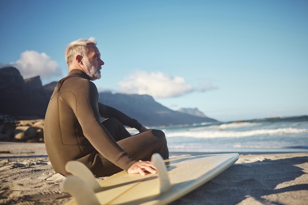 Tabla de surf y anciano en la playa para un viaje de aventura de surf esperando las olas del mar en la naturaleza para la actividad de deportes acuáticos Hombre en forma en el océano en verano para entrenamiento de surf en viajes en Sudáfrica