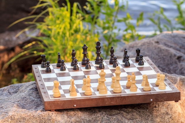 Tablero de ajedrez con piezas de ajedrez en roca con fondo de terraplén de río. Enfoque selectivo en piezas blancas. Juego de ajedrez al aire libre