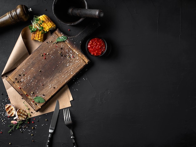 Tablero de madera pergamino tenedor cuchillo ajo a la parrilla brotes de maíz pimienta sal especias picador salsa mortero maja Fondo negro Vista superior