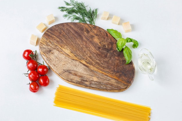 Tablero de madera, a su alrededor hay tomates, aceite de oliva, eneldo, queso, albahaca y pasta de espagueti.