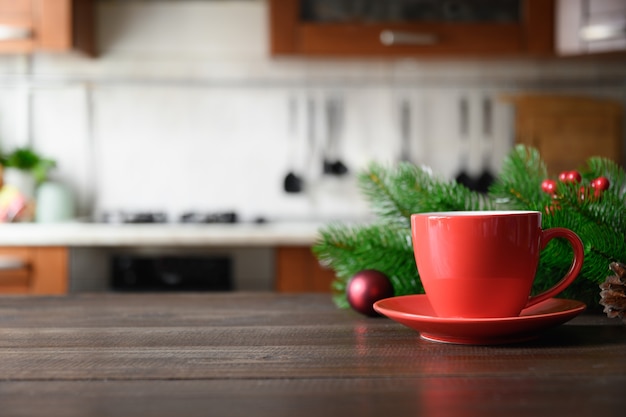 Tablero de madera con taza de café roja y cocina de Navidad borrosa.