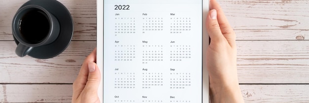 Tablet-Computer mit einer offenen App des Kalenders für das Jahr 2022 und einer Tasse Tee oder Kaffee in den Händen der Frau auf Holzhintergrund. Konzeptgeschäft oder To-Do-Listenziele. Ansicht von oben, flach liegend. Banner
