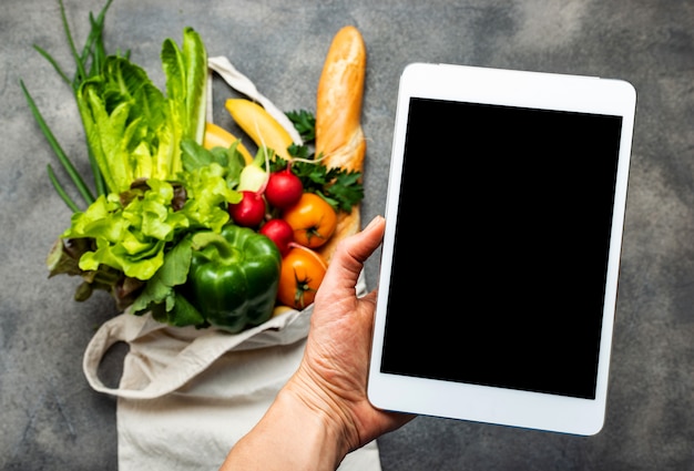 Tablet-Computer mit leerem Bildschirm in der weiblichen Hand über Einkaufstasche voll gesunder Lebensmittel.