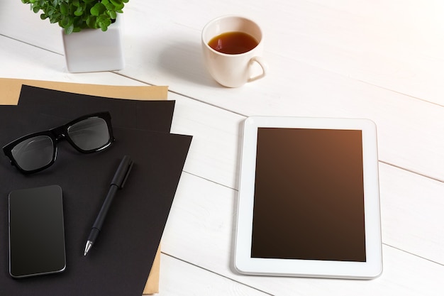 Tablet, folha em branco, copos e xícara de café na mesa de madeira do escritório. Fundo branco. Vista do topo. Copie o espaço. reflexo do sol