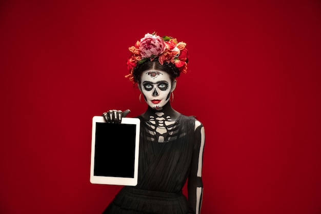 Tablet halten. Junges Mädchen wie Santa Muerte Saint Death oder Sugar Skull mit hellem Make-up. Porträt auf rotem Studiohintergrund isoliert. Feiern von Halloween oder Tag der Toten. Copyspace auf dem Bildschirm.