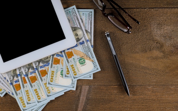 Tablet PC con un bolígrafo, gafas y billetes de dólar en una mesa de madera vista desde arriba.