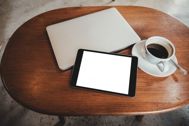 Tablet PC negro con pantalla blanca de escritorio en blanco con una computadora portátil y una taza de café en la mesa de madera