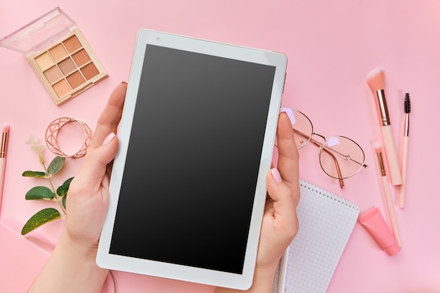 Tablet vazio nas mãos de uma mulher, óculos, canetas, acessórios de beleza, teclado, material de escritório, folha verde na superfície rosa