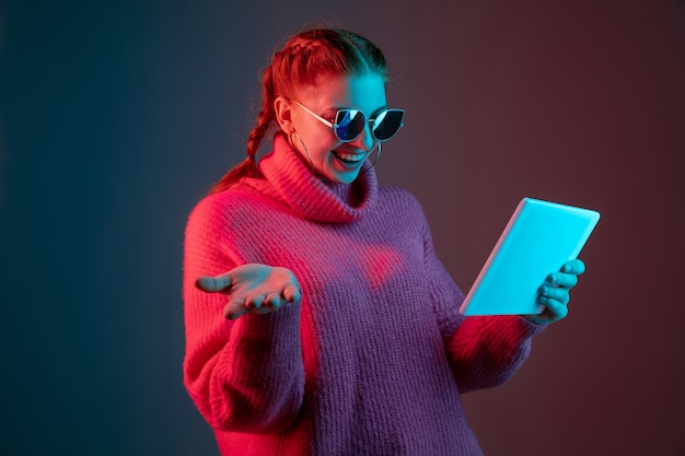 Tablet verwenden. Porträt der kaukasischen Frau auf Gradientenstudiohintergrund im Neonlicht isoliert. Schönes weibliches Modell mit Sonnenbrille, rotes Haar. Konzept der menschlichen Emotionen, Gesichtsausdruck, Anzeige.