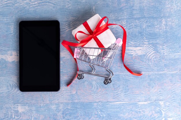 Tableta en blanco negro o pantalla de teléfono celular y carro con caja de regalo sobre fondo de madera azul, espacio de copia. Concepto de felicitaciones en línea. Concepto de compra online