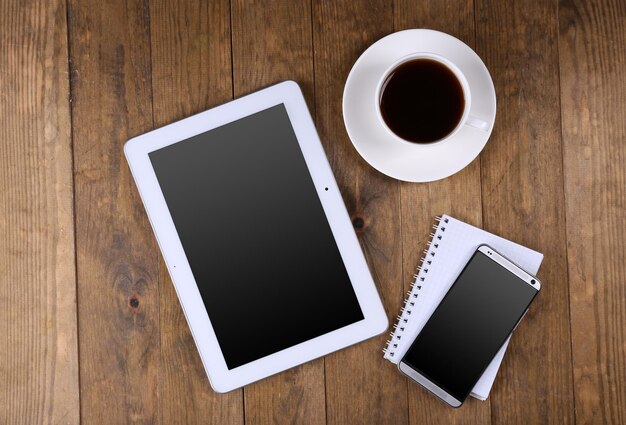 Tableta en blanco y teléfono móvil con cuaderno y taza de café sobre fondo de madera