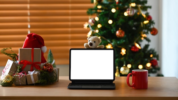 Tableta de computadora, taza de café y cajas de regalo en la mesa de madera cerca del árbol de Navidad en casa.