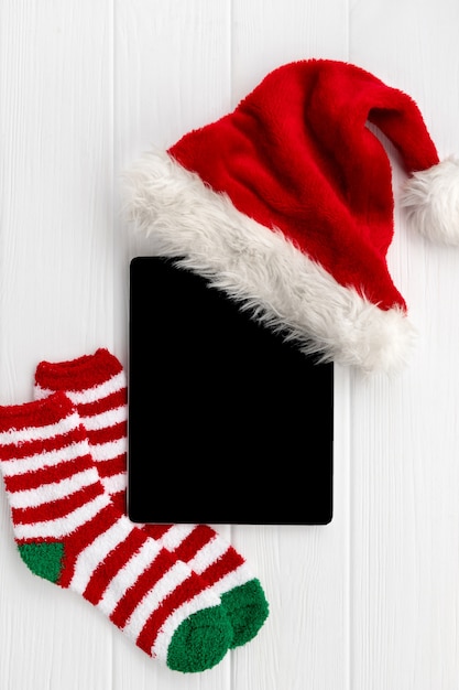 Tableta digital con gorro de Papá Noel y calcetines en madera blanca. Plano de navidad