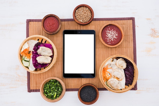 Tableta digital de pantalla blanca en blanco envolvente con salsa; Cebollino y semillas de sésamo sobre mantel sobre fondo de textura
