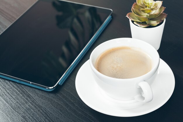 Tableta digital con pantalla negra, taza de café y pequeña planta en una mesa de trabajo