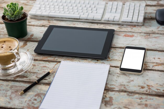 Tableta digital con teclado de computadora, teléfono inteligente, planta de maceta, bolígrafo, bloc de notas y taza de café