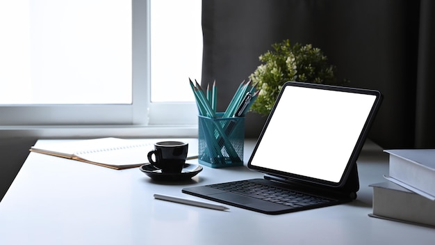Tableta digital con teclado inalámbrico, taza de café de planta en maceta y papelería en mesa blanca