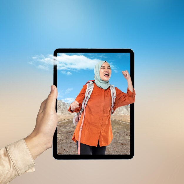 Una tableta de la mano humana con una vista de pantalla de una chica asiática en una bufanda con una mochila de pie con colinas y una vista de acantilado de roca con cielo azul