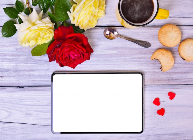 Tableta con pantalla blanca, taza de café negro y ramo de rosas.