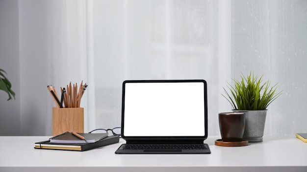 Tableta con pantalla en blanco, portalápices, planta de la casa, taza de café y cuaderno sobre mesa blanca.