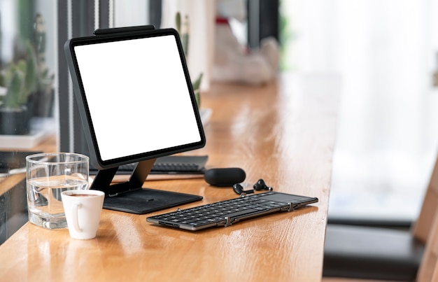 Tableta de pantalla en blanco en el soporte con teclado y gadget en la mesa de mostrador de madera en la cafetería.