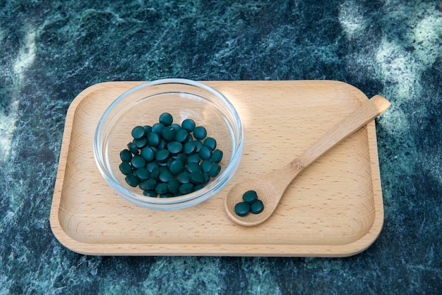 tabletas de espirulina en un recipiente de vidrio y una cuchara de madera en una bandeja de madera sobre un fondo de mármol verde