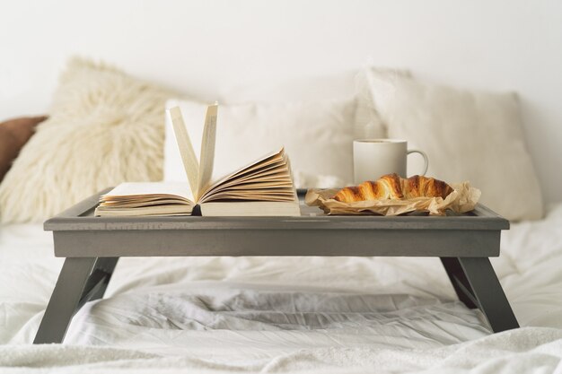 Tablett mit Croissant, Tasse Kaffee und Buch auf weißem, sauberem Bett.