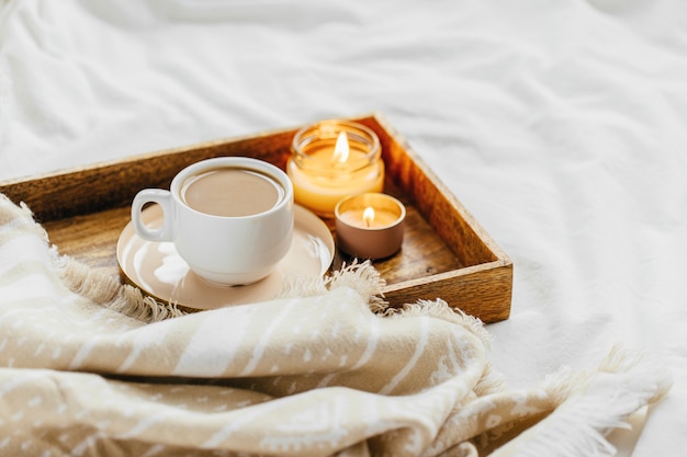 Tablett mit Kaffee und Kerzen mit warmem Plaid auf weißer Bettwäsche. Frühstück im Bett.