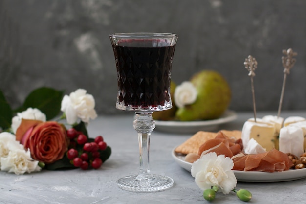 Taça de cristal com vinho tinto no meio da moldura, ao lado de prato com presunto e queijo camembert e flores