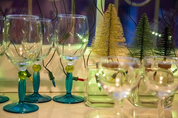 taças de vidro com pernas azuis ficam em uma mesa branca perto da janela ao lado de pequenas árvores de Natal decorativas. fechar-se. conceito de noite aconchegante