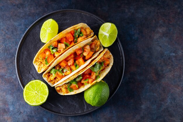 Taco mexicano e limas em um fundo escuro. Tacos com carne de porco picada, tomate e ervas. Taco com carne e legumes em um prato preto. Copie o espaço. Vista do topo