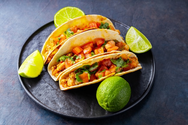 Taco mexicano e limas em um fundo escuro. Tacos com carne de porco picada, tomate e ervas. Taco com carne e legumes em um prato preto. Copie o espaço. Vista do topo