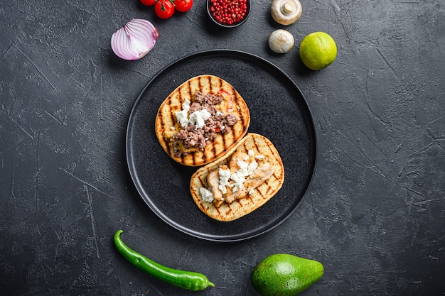 Taco mexicano tradicional con pollo y ternera con ingredientes al lado de la placa negra sobre fondo negro con textura vista superior.