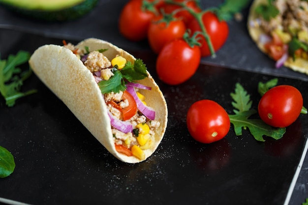 Tacos mexicanos con carne de cerdo, verduras y especias en una placa de piedra negra sobre un fondo oscuro con ingredientes para tacos