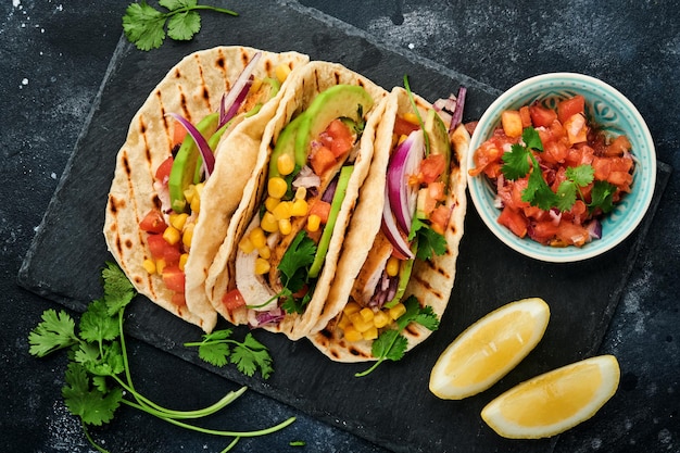 Tacos mexicanos com frango grelhado, abacate, grãos de milho, tomate, cebola, coentro e salsa na mesa de pedra preta. Comida de rua tradicional mexicana e latino-americana. Vista do topo.