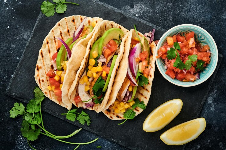 Tacos mexicanos com frango grelhado, abacate, grãos de milho, tomate, cebola, coentro e salsa na mesa de pedra preta. Comida de rua tradicional mexicana e latino-americana. Vista do topo.