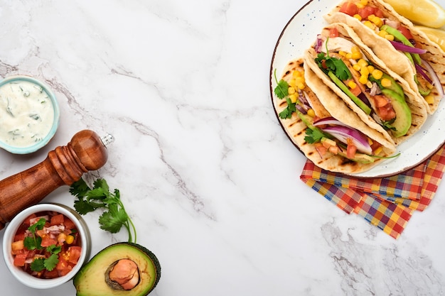 Tacos mexicanos con pollo a la parrilla, aguacate, granos de elote, tomate, cebolla, cilantro y salsa en la mesa de piedra blanca. Comida callejera tradicional mexicana y latinoamericana. Vista superior.