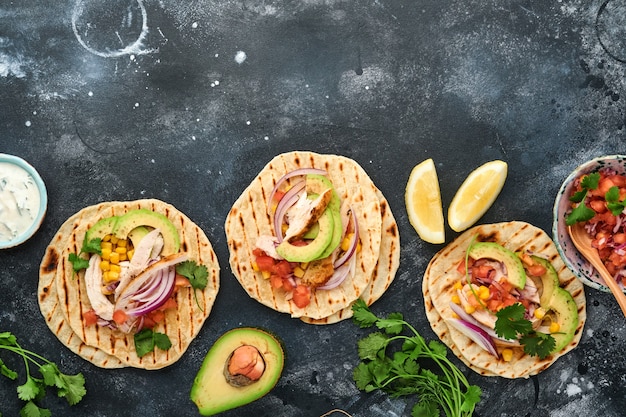 Tacos mexicanos con pollo a la parrilla, aguacate, granos de maíz, tomate, cebolla, cilantro y salsa en la mesa de piedra negra. Comida callejera tradicional mexicana y latinoamericana. Vista superior.