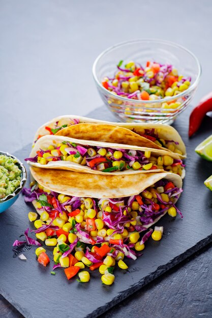 Tacos vegetarianos con diversas verduras, guacamole y rodajas de limón sobre fondo oscuro. Tacos de maíz dulce, repollo morado y pimiento en una tabla de pizarra. Copia espacio