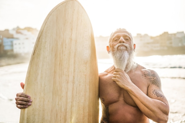 Tätowierter Senior Surfer mit Vintage Surfbrett am Strand bei Sonnenuntergang - Fokus auf Gesicht