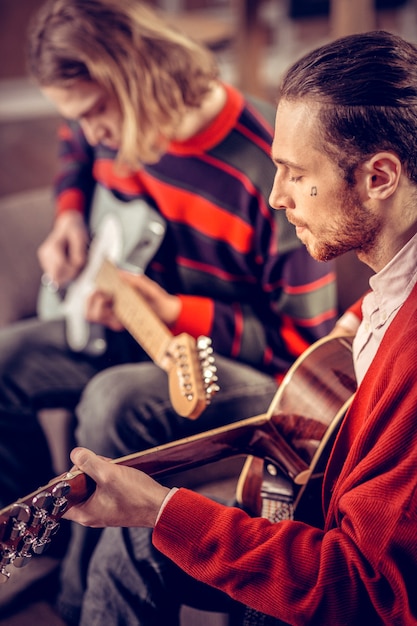 Tätowierung im Gesicht. Bärtiger, dunkelhaariger Musiker mit kleinem Tattoo im Gesicht, der Gitarre spielt