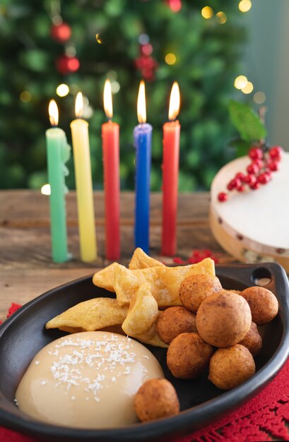 Tag bei Kerzenschein. Typisch kolumbianisches Essen. Buñuelo, Vanillepudding und Flocken mit Kerzen und Weihnachtsbaum im Hintergrund. Traditioneller kolumbianischer Feiertag. Vertikales Video.