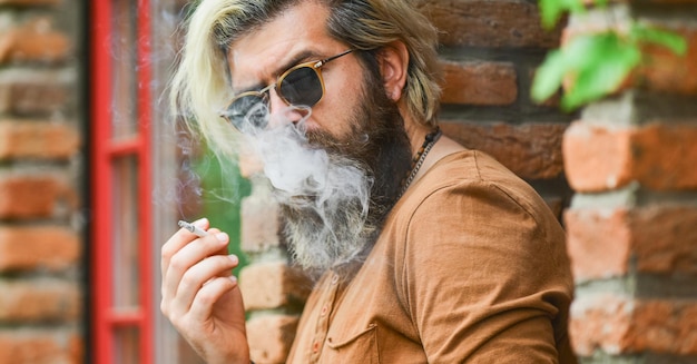 Tag in Rauch. eine Rauchpause machen. Hübscher stilvoller Mann, der draußen in der städtischen Umgebung raucht. Hipster-Lifestyle. Mann raucht eine Zigarette. Geschäftsmann in Gläsern, die Zigarette auf der Straße rauchen.