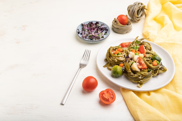 Tagliatelle grüne Spinatnudeln mit Tomaten-, Erbsen- und Mikrogrünsprossen auf einem weißen Holztisch und gelbem Textil. Seitenansicht, Kopierraum.