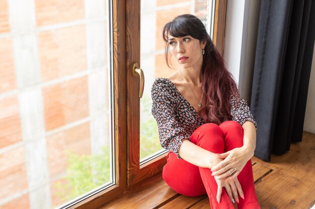 Tagträumen, Nachdenklichkeit und Einsamkeit Konzept - Junge kaukasische Frau, die in ihrem Wohnzimmer sitzt und durch großes Fenster schaut.