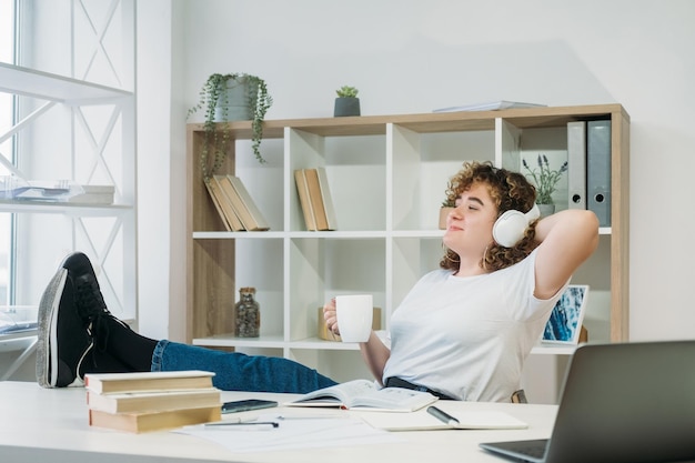 Tagträumendes Mädchen Stressabbau Tag der Harmonie Sorgloses Wochenende Ruhige, entspannte, übergewichtige Frau mit lockigem Haar, die Musik in Kopfhörern mit Kaffeetasse in einer modernen, hellen Wohnung genießt