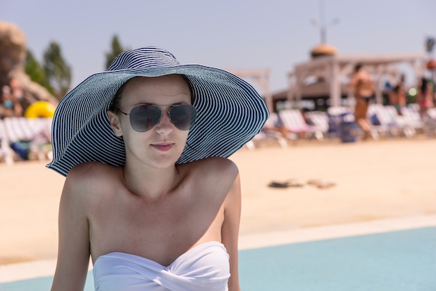 Taille bis Nahaufnahme einer jungen Frau im Urlaub mit Sonnenhut, Sonnenbrille und weißem Bikini beim Sitzen auf der sonnigen Terrasse des öffentlichen Resort-Swimmingpools