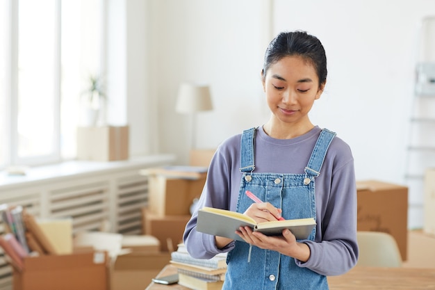 Taille hoch Porträt der jungen asiatischen Frau, die im Planer schreibt, während sie im neuen Haus oder in der neuen Wohnung mit Pappkartons steht