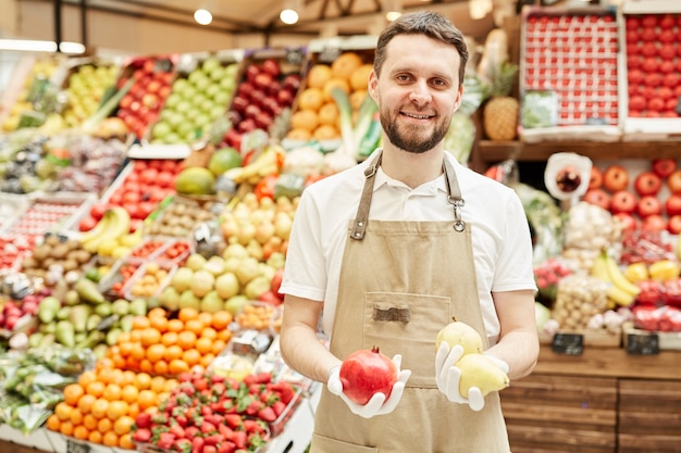 Taille hoch Porträt des bärtigen Mannes, der Schürze trägt und lächelt, während frisches Obst und Gemüse am Bauernmarkt verkauft